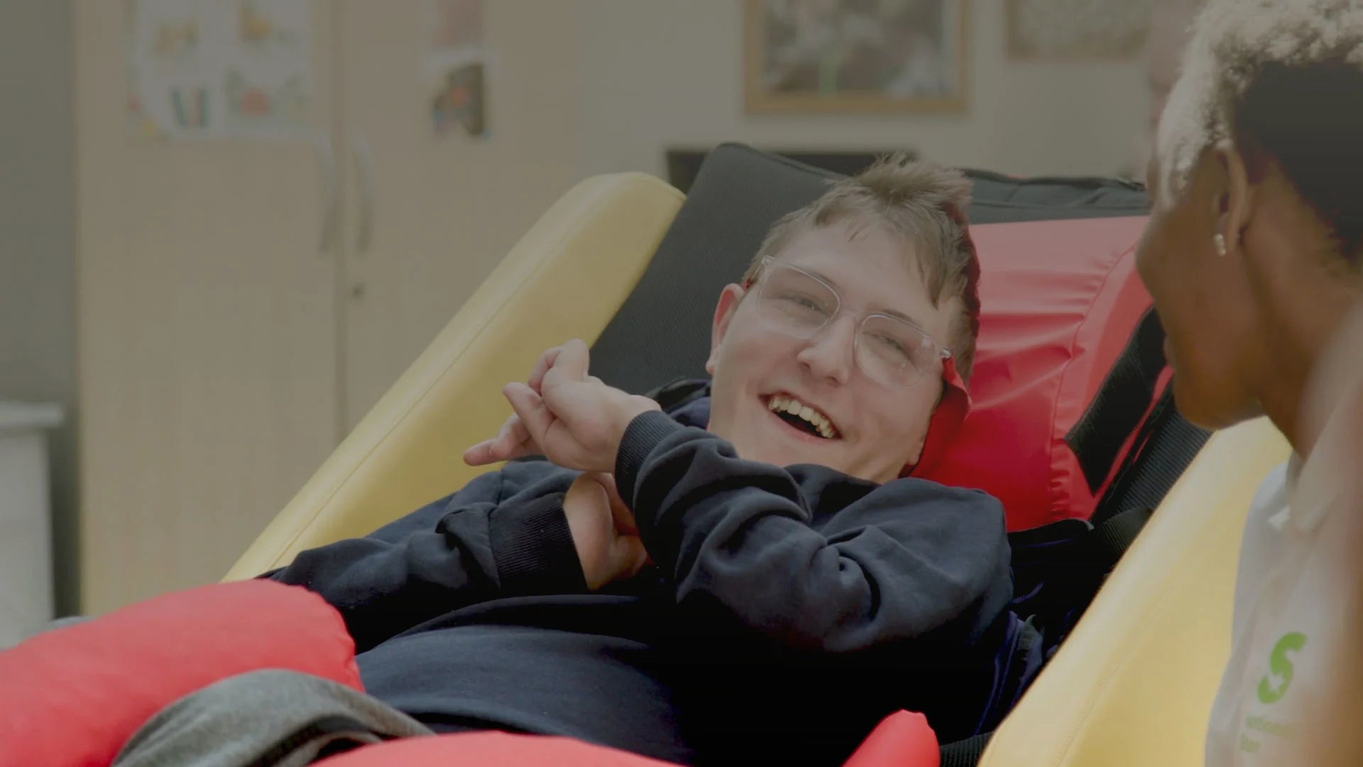 Person lying on a colourful acheeva chair with another person beside them in an indoor setting.