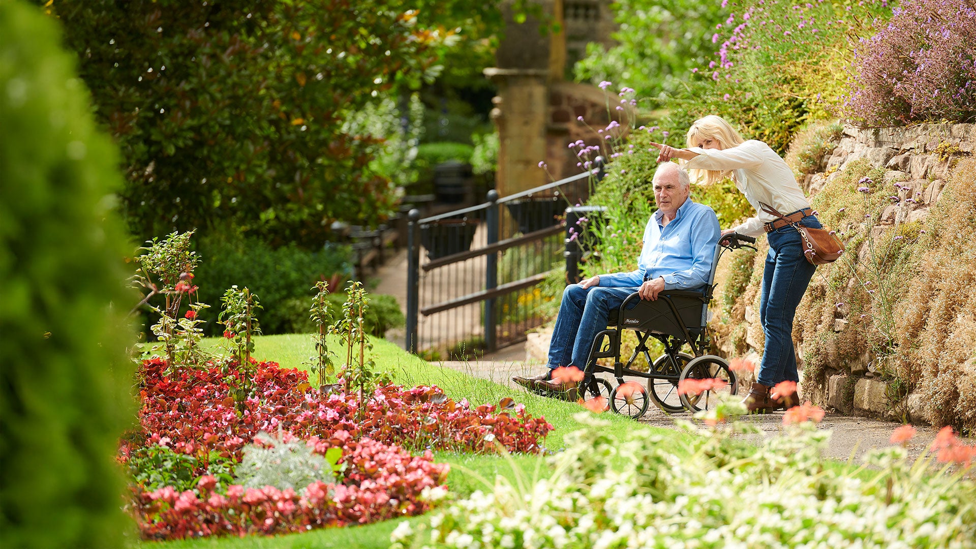 Man in a wheelchair being pushed by a woman in a garden with flowers and a stone wall.