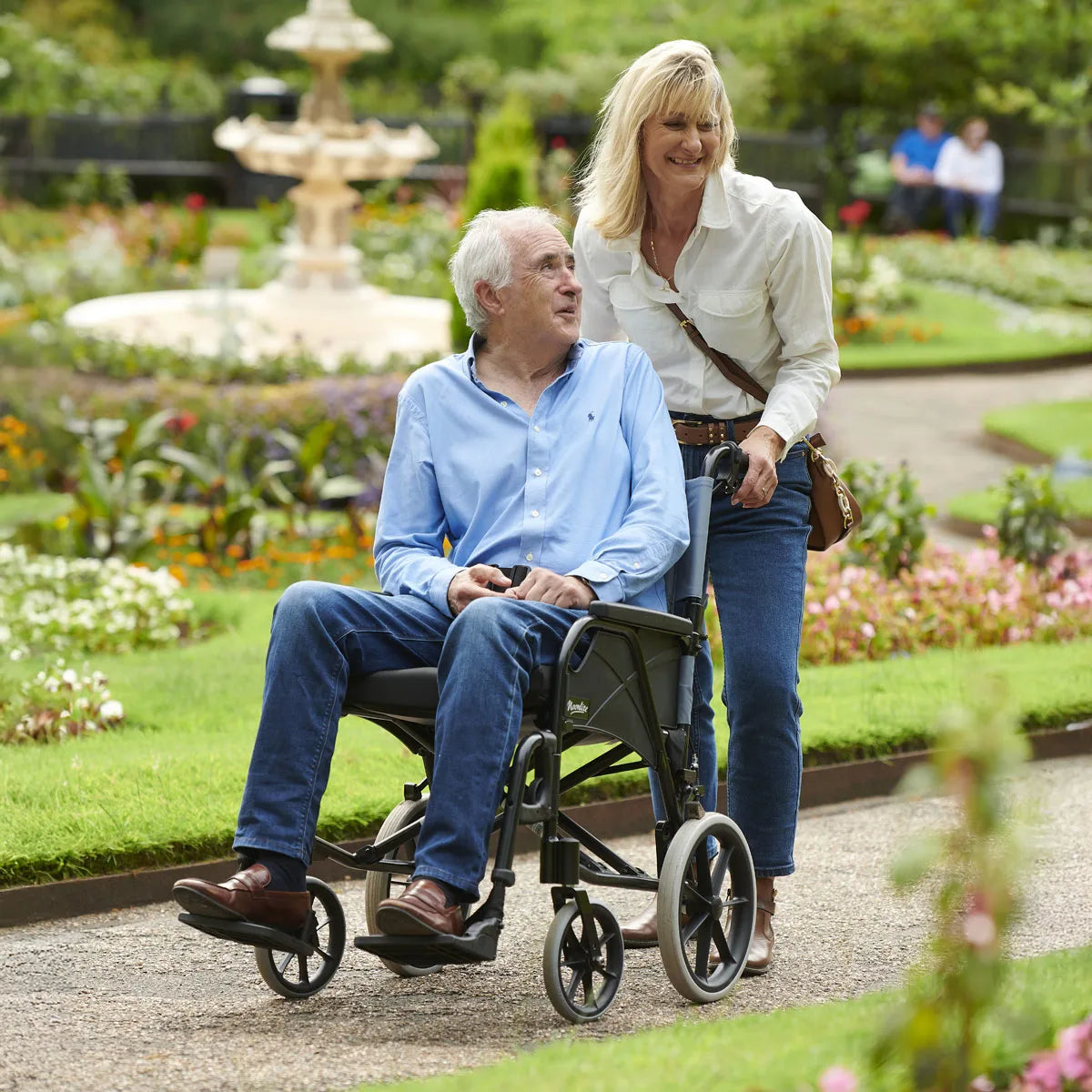An elderly man in a wheelchair being pushed by a woman in a garden path.