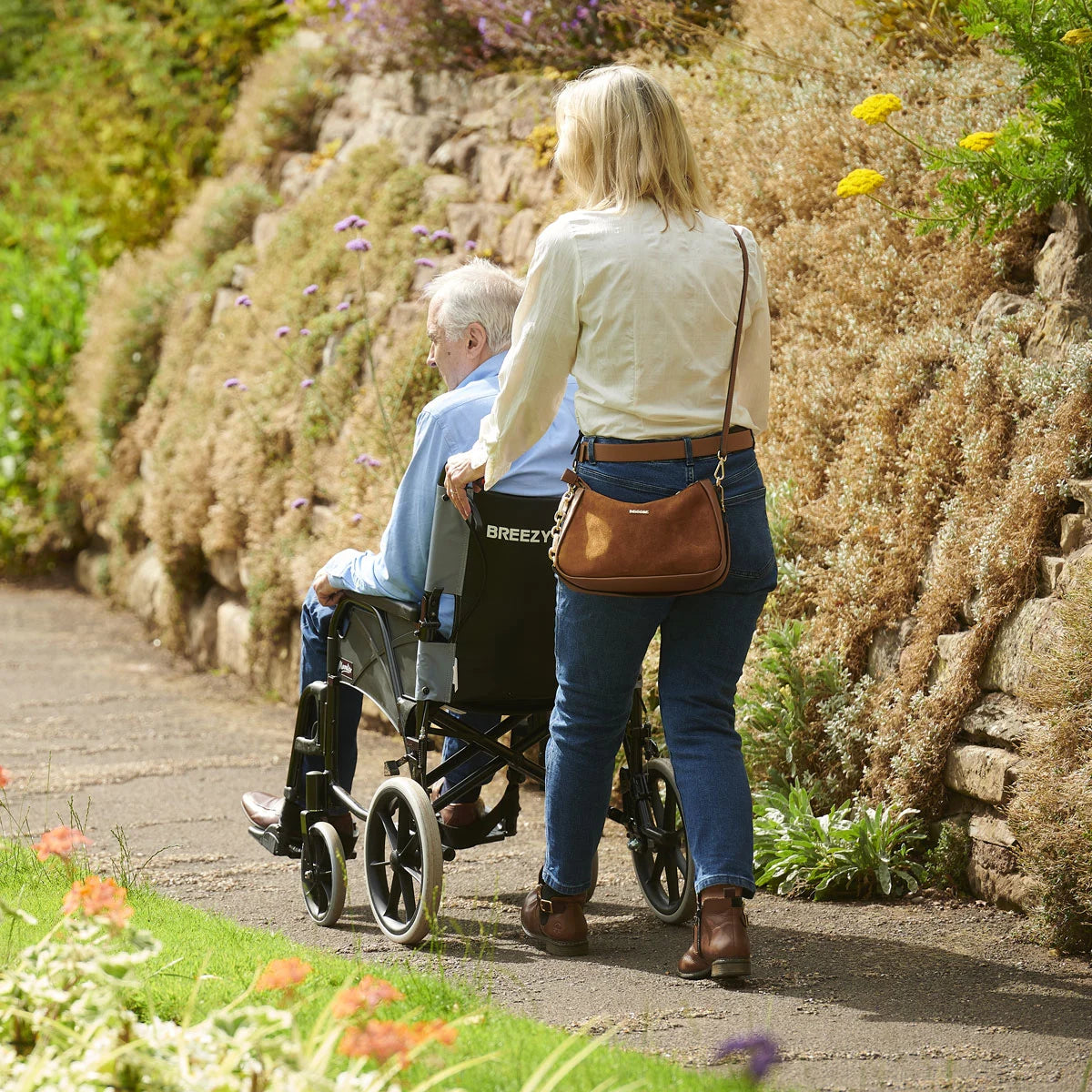 Person pushing a wheelchair with an elderly person along a garden path.