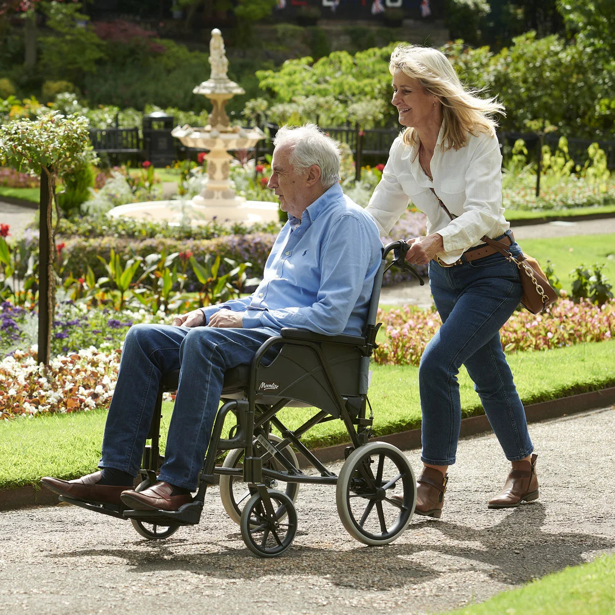An elderly man in a wheelchair being pushed by a woman in a park with a fountain and garden in the background.