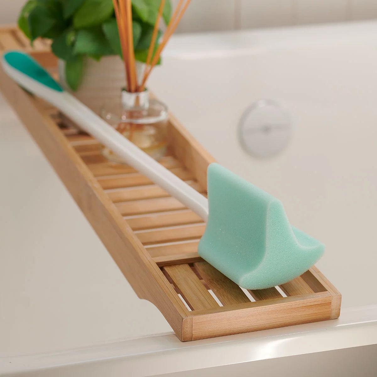 Bamboo bath caddy with green toe and foot sponge and white brush on a bathtub ledge.