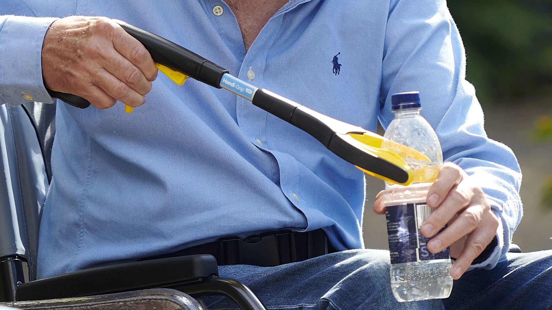 Person in a wheelchair using the mini reacher to pick up a bottle, with a blurred background