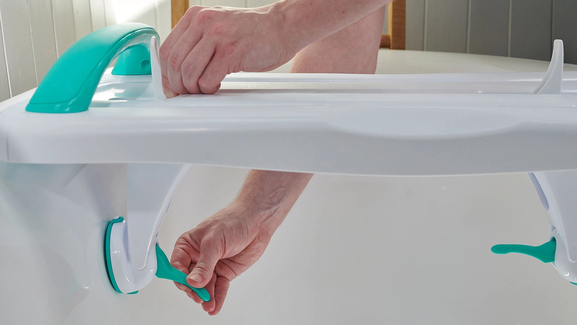 Person adjusting a white bathtub support device with green handles in a bathroom setting.