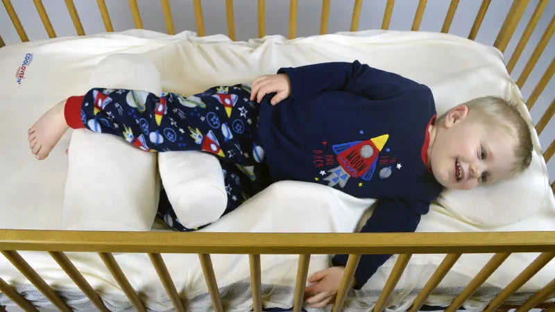 Child in pyjamas lying in a crib with a white mattress and supportive pillows