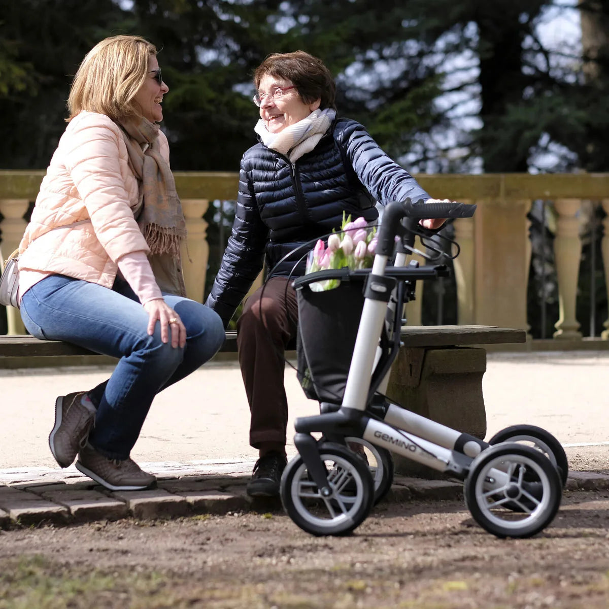 Two people are sat on a bench with a rollator that has a black bag with pink flowers inside.