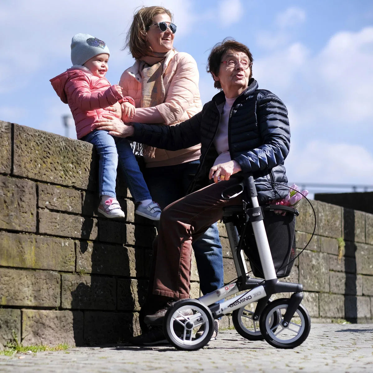 Two adults and a child are sat alongside a all with one person holding a rollator with a black bag and pink flowers inside