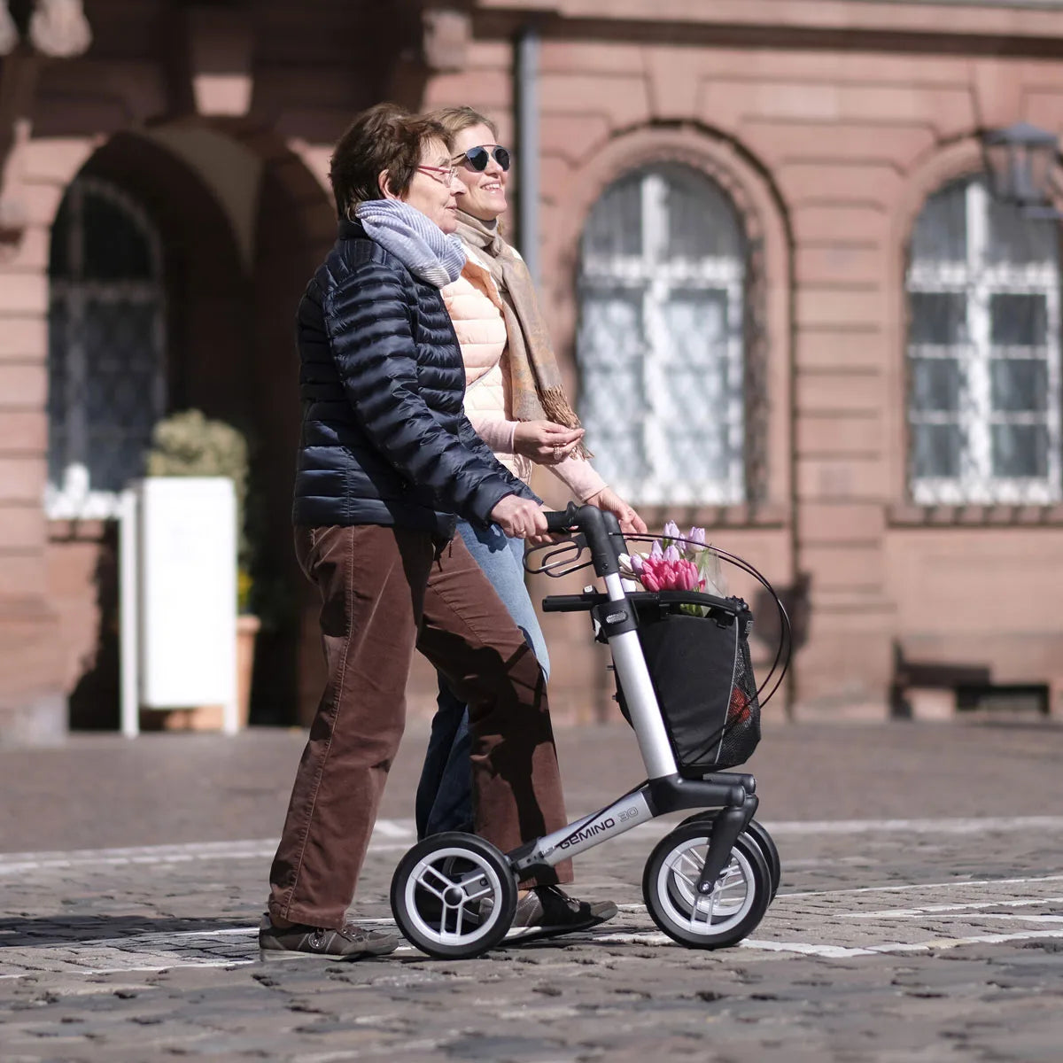 Two people with a rollator that has a black bag with pink flowers inside on a cobblestone street in front of a building.