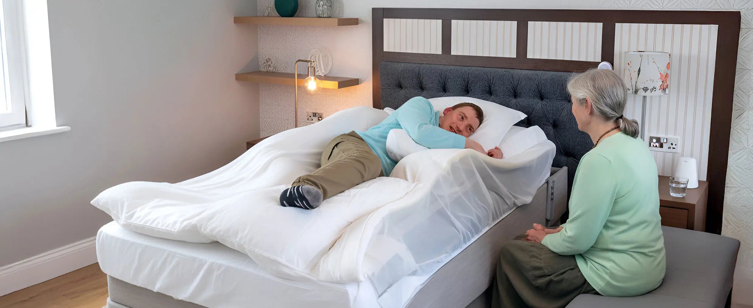 Man lying on a Symmetrisleep sleep positioning system, with woman sitting next to him in a bedroom setting.