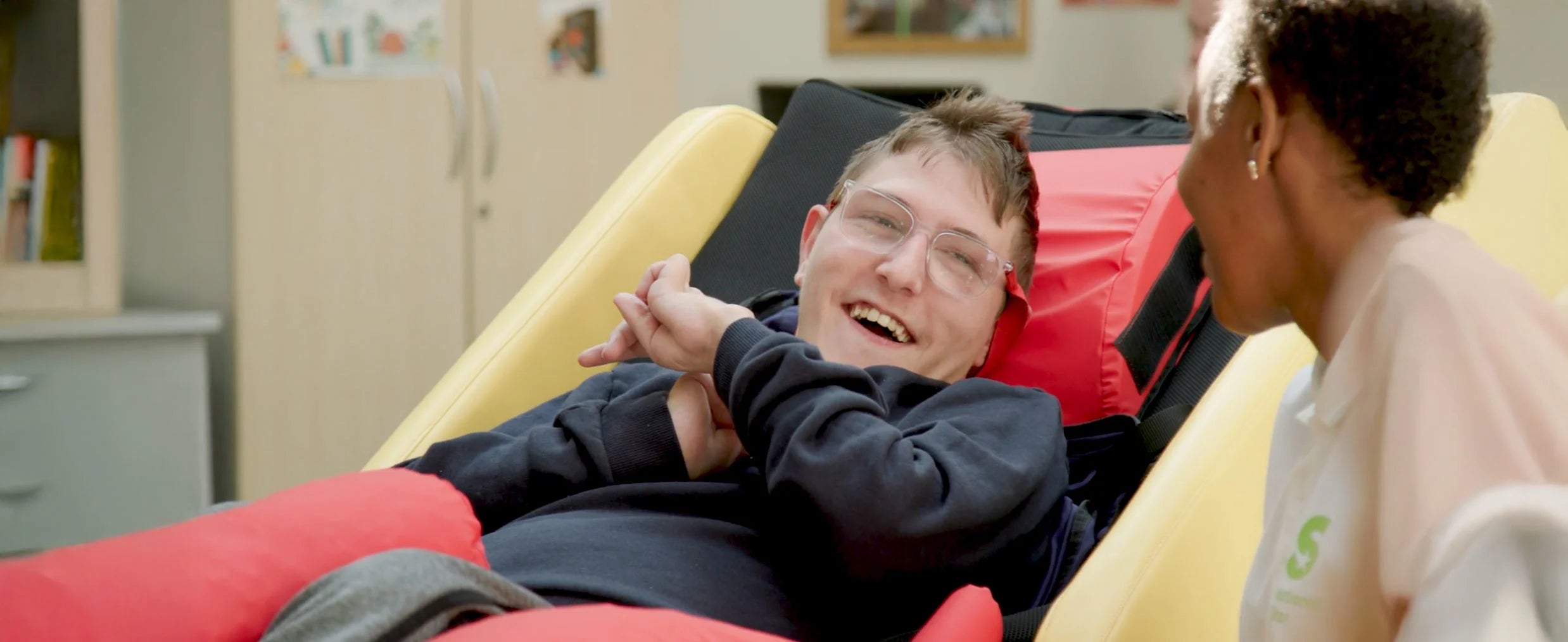 Person lying on a red cushioned acheeva with another person sitting next to them, in a room.