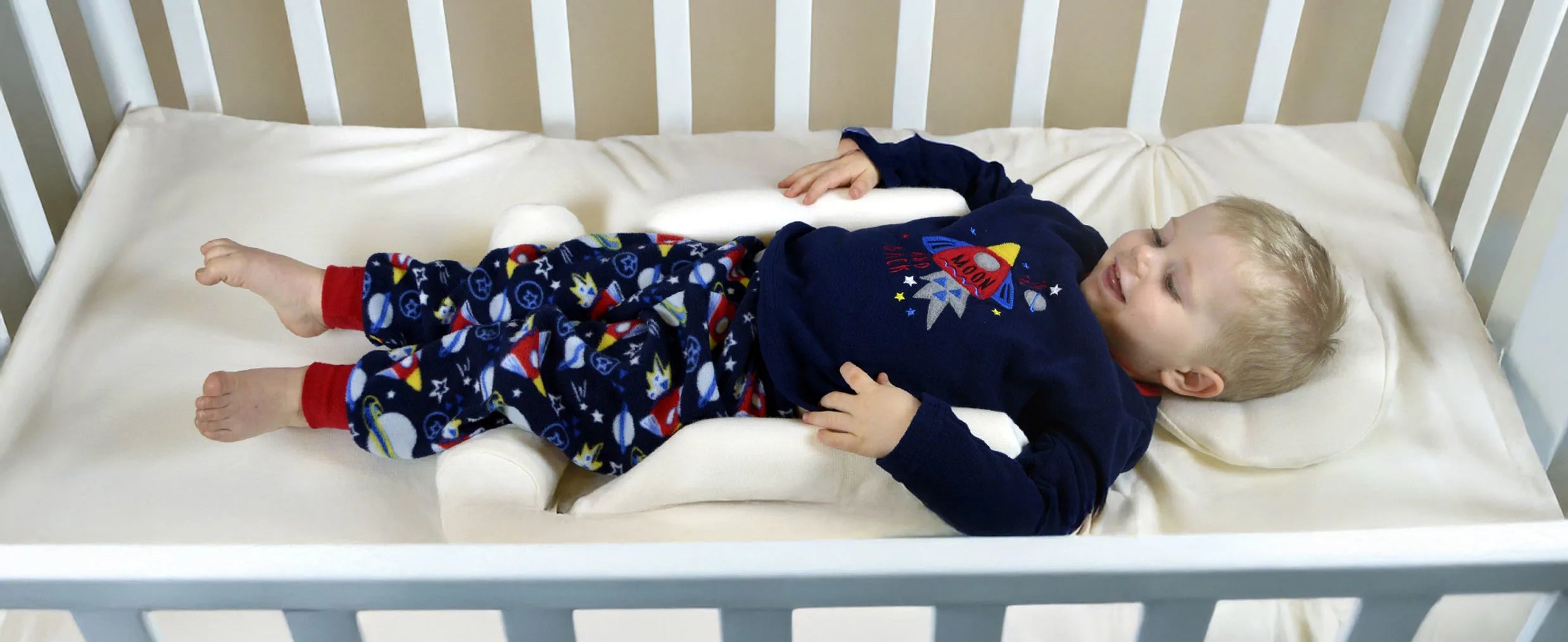 Child lying in a crib wearing a navy blue outfit with space-themed using supportive pillows.