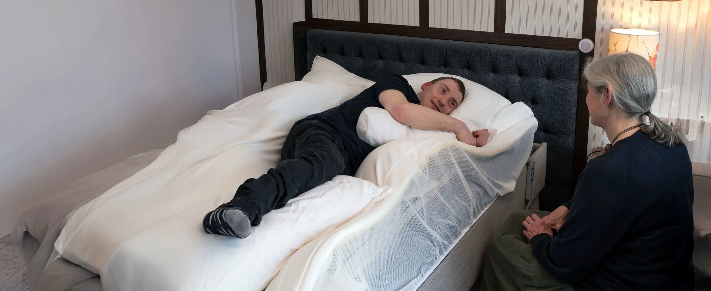 Person lying on a bed with supportive pillows and another person sitting beside them, in a bedroom setting.