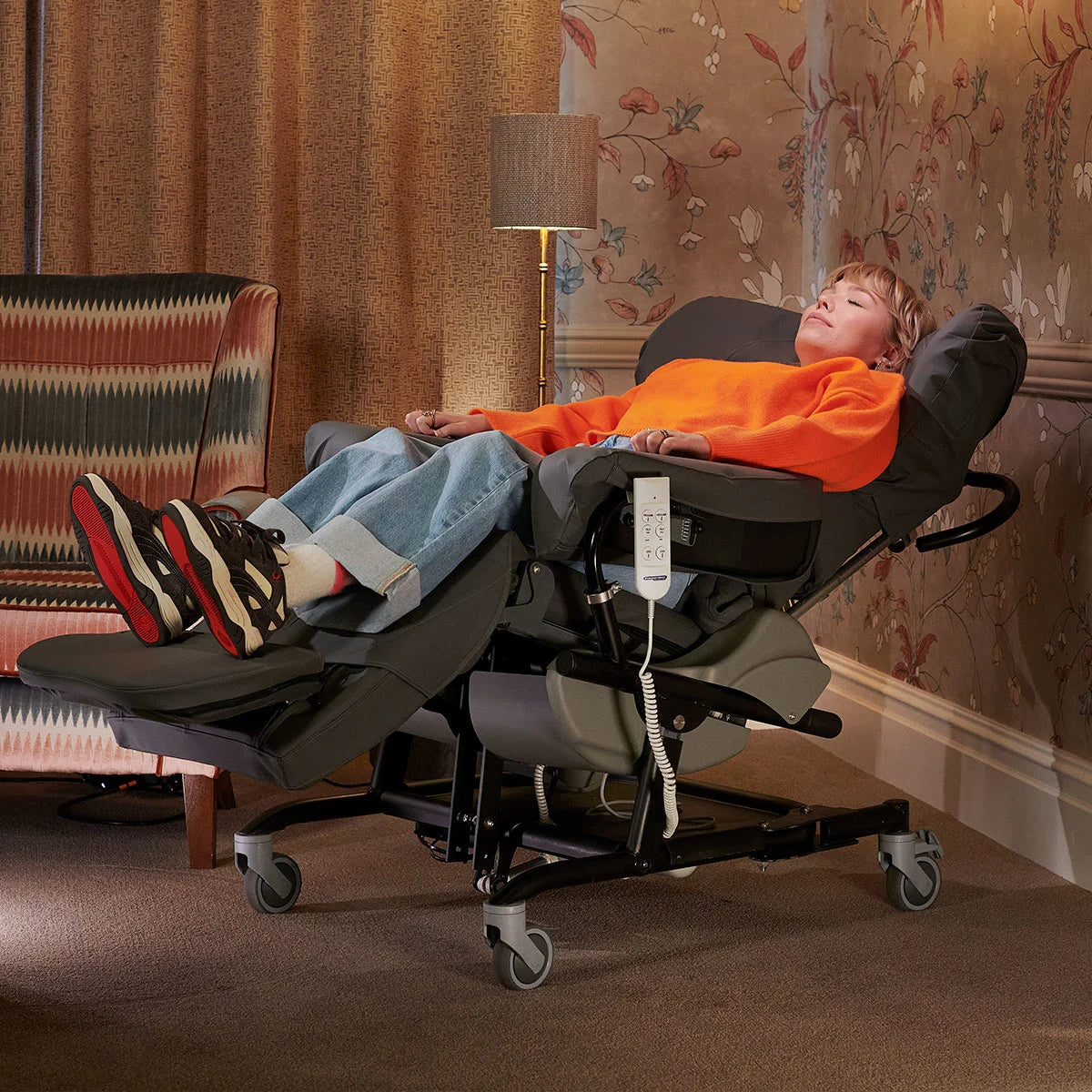 A woman relaxing in a grey reclining chair wearing an orange jumper ad blue jeans with a lamp and wallpaper in the background