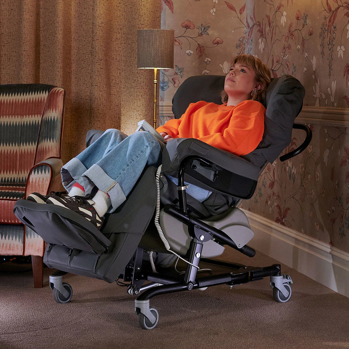a woman lying down in a reclining grey chair wearing an orange jumper and blue jeans holding a book with a lamp and colourful wallpaper in the background