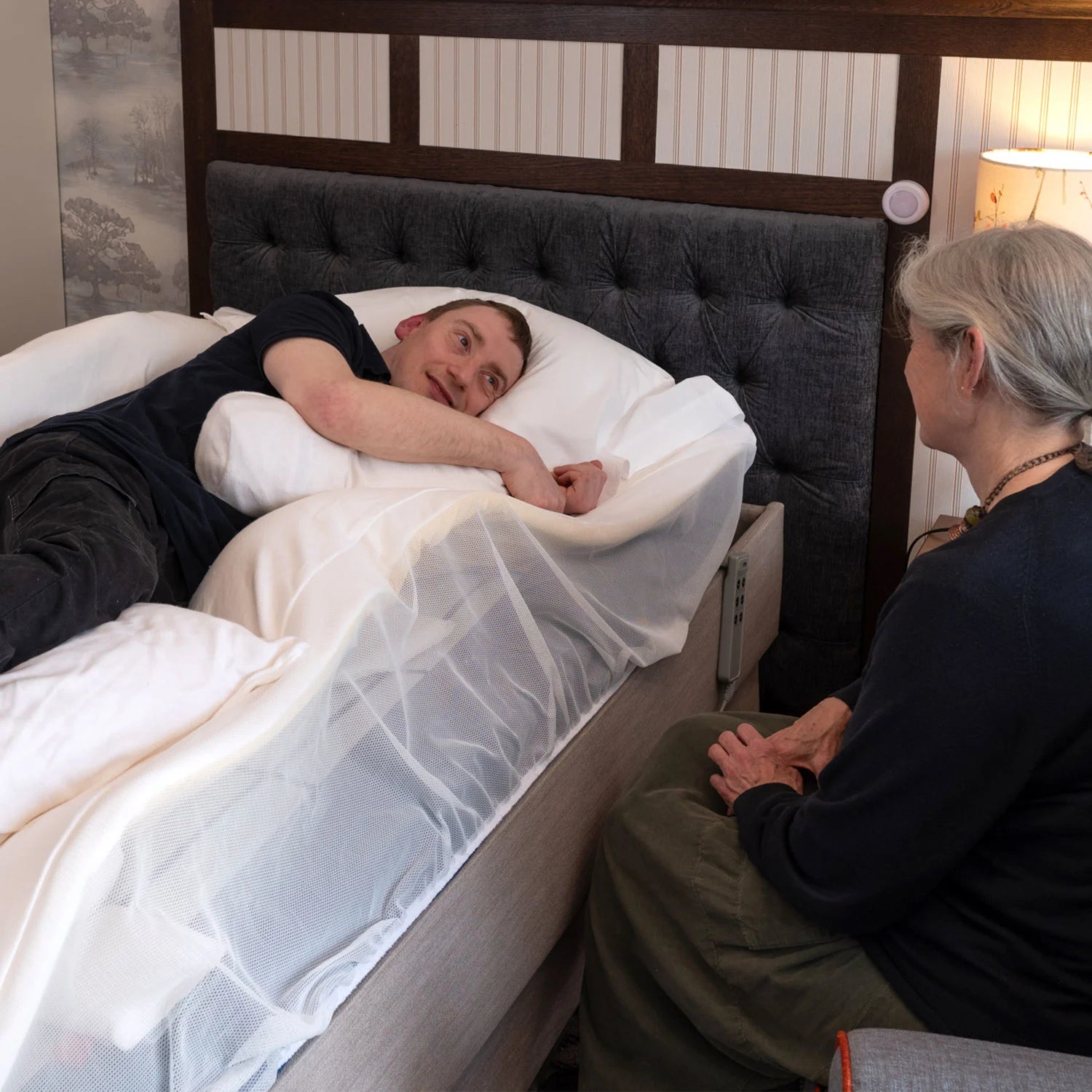 Person lying in bed with supportive pillows, interacting with another person.