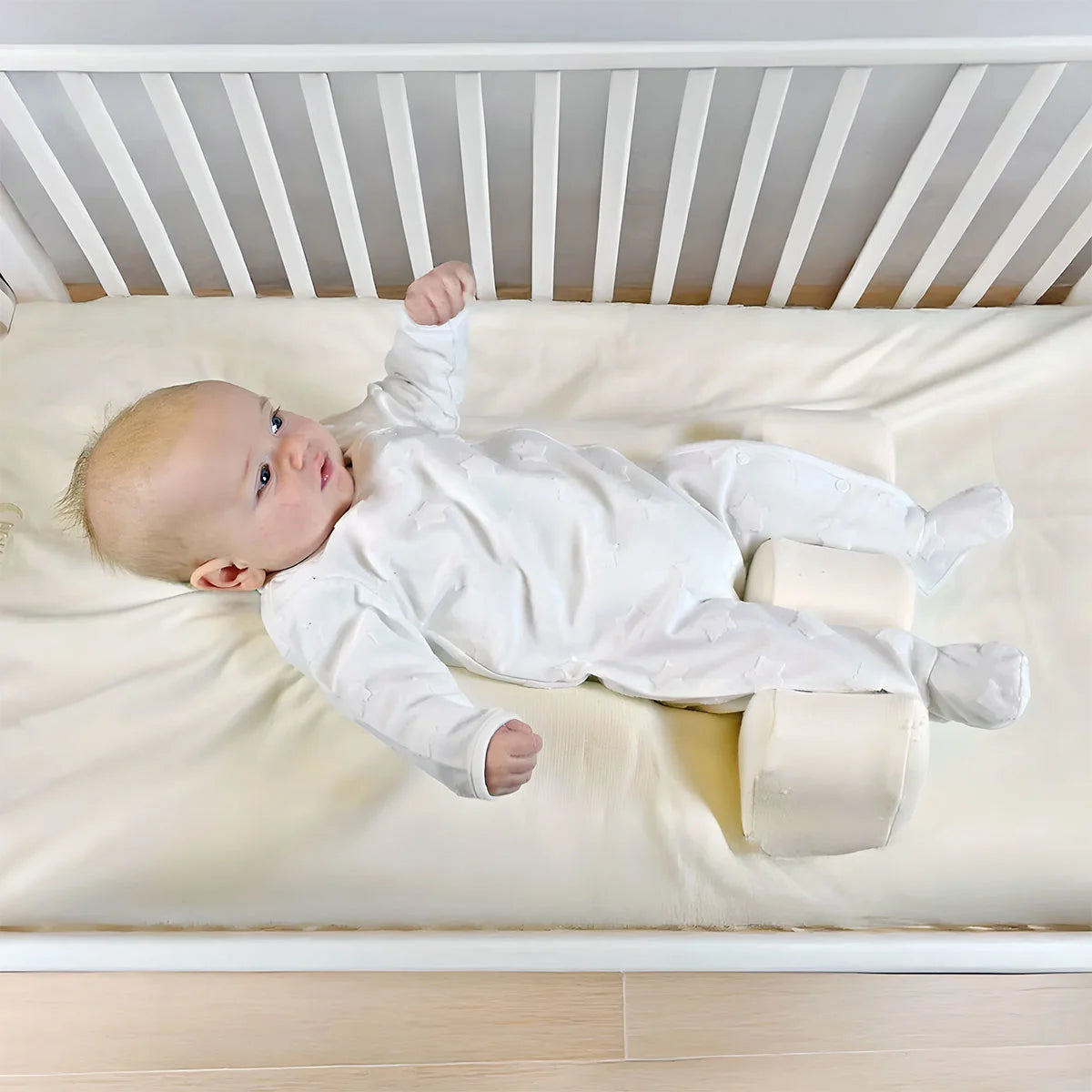 Baby lying on a white crib mattress with a white crib in the background with their legs between supportive pillows.