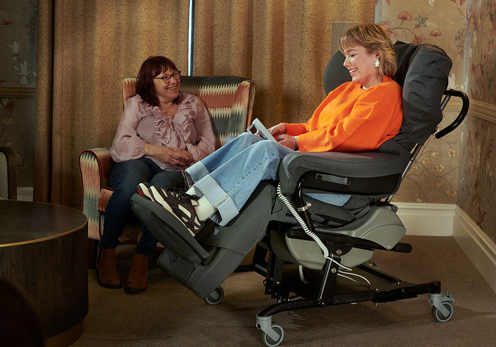 Woman in an orange sweater using a tablet in a reclining chair, with another woman sitting in a chair in the background.