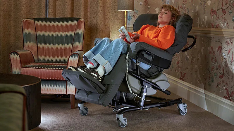 Person using a reclining chair in a home setting with a patterned armchair and floral wallpaper.