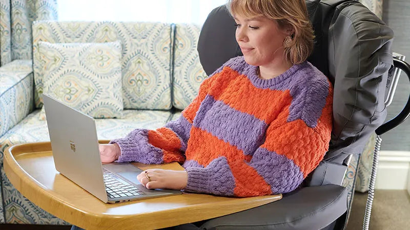 Person wearing a colorful sweater in a reclining chair using a laptop on a small table.