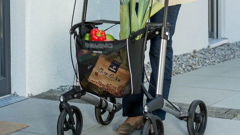Person walking with groceries in basket of Gemino 30 Rollator.  
