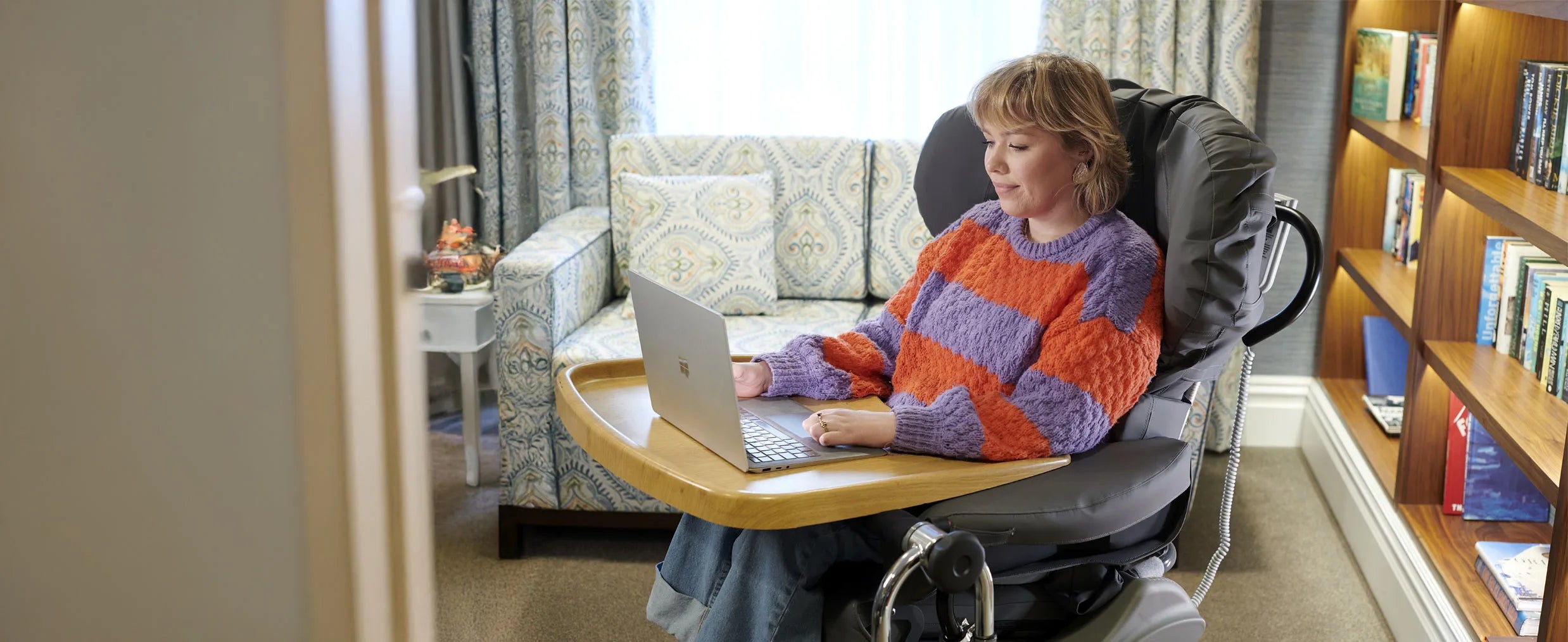 Person using a laptop sat in a symmetry sleep with a bookshelf and sofa in the background.