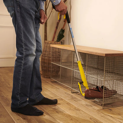 Person using a yellow and black shoe helper grabber to get their shoes on a wooden floor next to a metal wire basket.