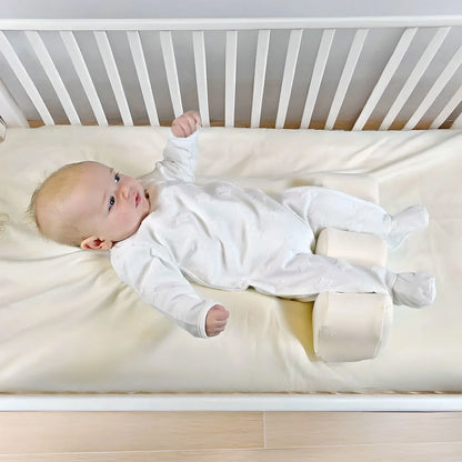 Baby lying on a white crib mattress with a white crib in the background with their legs between supportive pillows.