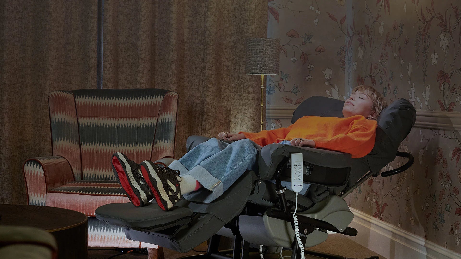 Person lying in a recliner chair with a remote control, in a room with patterned wallpaper and a striped armchair.